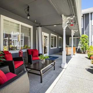 A cozy patio area with wicker furniture and red cushions, surrounded by plants and shaded under a covered structure.