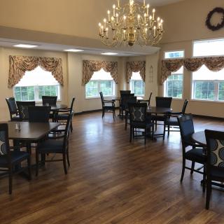 A dining area with several tables and chairs, featuring a chandelier and large windows with decorative curtains.