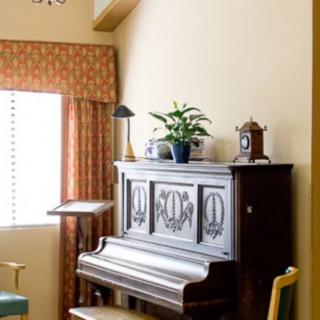 A cozy room interior featuring an upright piano adorned with a plant and a lamp, complemented by a nearby chair and window with curtains.