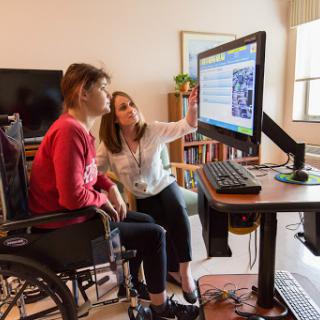 A healthcare professional assists a person in a wheelchair with technology at a desk.