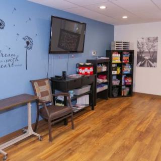 A cozy room with a wall-mounted television, shelves stocked with various items, and a comfortable chair on a hardwood floor.