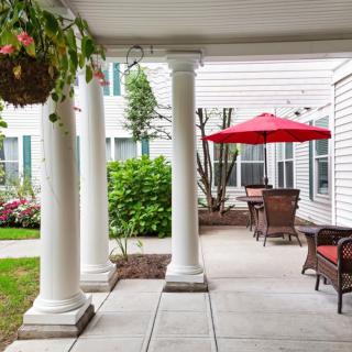 A peaceful porch with columns and outdoor seating under a red umbrella, surrounded by lush greenery.