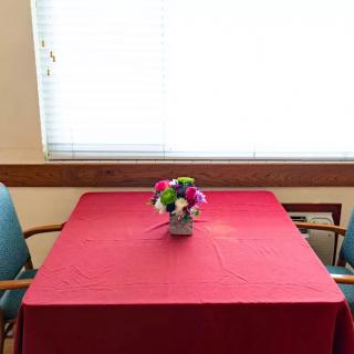 A dining table with a red tablecloth and a centerpiece of colorful artificial flowers, surrounded by chairs next to a window.