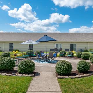 A well-maintained outdoor patio area with tables, chairs, and umbrellas surrounded by neatly trimmed bushes under a sunny blue sky.