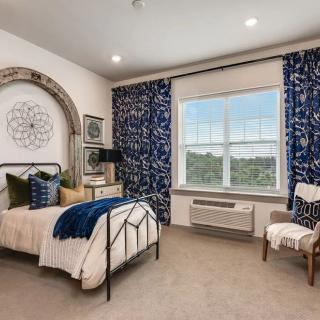 A well-decorated bedroom featuring a bed with blue and white accents, a large window with patterned curtains, and stylish furniture.