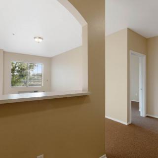 An empty apartment interior with beige walls, a carpeted floor, and an archway leading to another room.