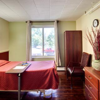 A bright, simple hospital room featuring a red bedspread and wooden furniture.