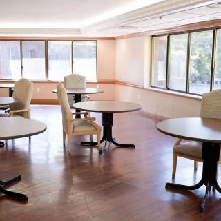 An empty room with tables and chairs, well-lit by natural light coming through large windows.