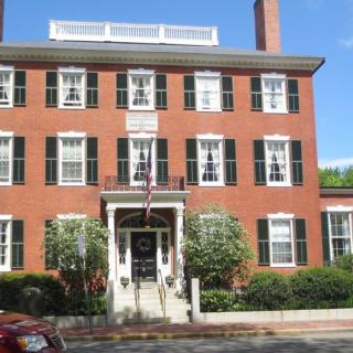 A historic brick building with green shutters and a central entrance, surrounded by plants.
