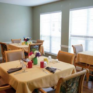 A cozy dining room with tables set for a meal, featuring natural light from large windows.