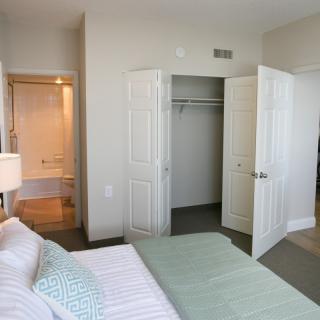 A modern, minimalist bedroom with a view into an adjacent bathroom; features include a neatly made bed and open closet doors.