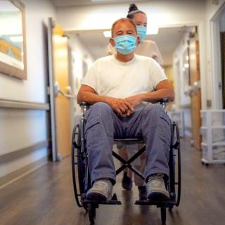 A nurse pushes a patient in a wheelchair down a hospital corridor, both wearing masks.
