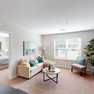 A bright and cozy living room with a beige sofa, colorful pillows, and a round coffee table near the window.