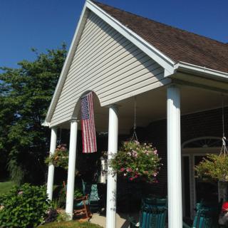 A charming house with a porch featuring white columns, hanging plants, and an American flag on a sunny day.