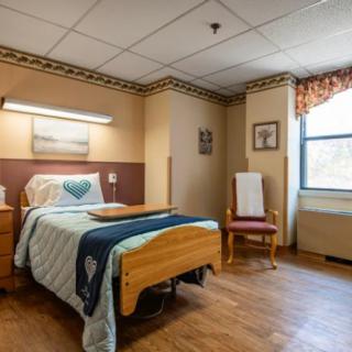 A cozy hospital room with a single bed and wooden furniture, featuring a window and a comfortable chair.