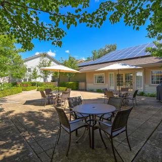 A sunny outdoor patio with tables and chairs, surrounded by trees and near a building.