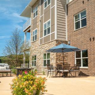 A sunny outdoor patio area with tables, chairs, and an umbrella beside a brick building.