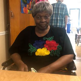 An elderly woman in a colorful shirt smiles while seated in a wheelchair, enjoying her time indoors.