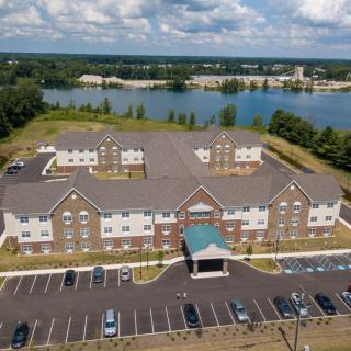 Aerial view of a large building with a parking lot, set against a scenic backdrop of a lake and cloudy sky.
