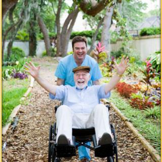 An elderly man in a wheelchair being pushed by a caregiver, both smiling and enjoying time outdoors in a garden.