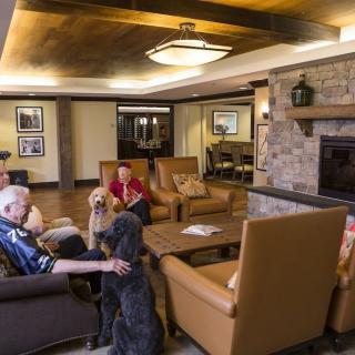 Senior residents relaxing in a communal lounge with dogs by the fireplace.