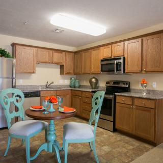 A modern kitchen with wooden cabinets and stainless steel appliances, featuring a small dining table with decorative chairs.