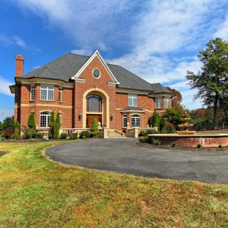 A large brick luxury mansion with a circular driveway and landscaped front yard under a partly cloudy sky.