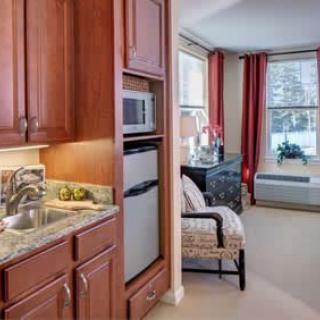 A cozy living space featuring a kitchenette with wooden cabinetry, equipped with a microwave and refrigerator, alongside an adjoining seating area with an armchair by the window.