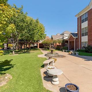 A scenic outdoor area with benches and a pathway alongside a brick building, surrounded by trees and greenery.