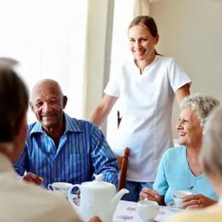 A caregiver interacts with elderly individuals around a table, sharing smiles and conversation.