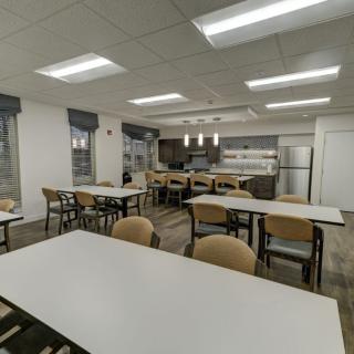 A modern cafeteria with neatly arranged tables and chairs, featuring a kitchen area in the background.