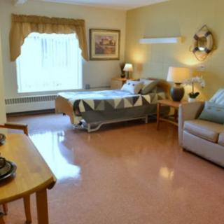 A cozy and well-lit bedroom in a nursing home or hospital setting, featuring a single bed, sofa, and various lamps.