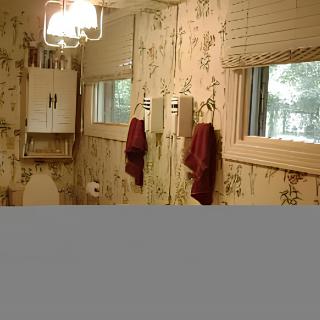 A vintage-style bathroom featuring floral wallpaper, a mirror, red towels, and a window with blinds.