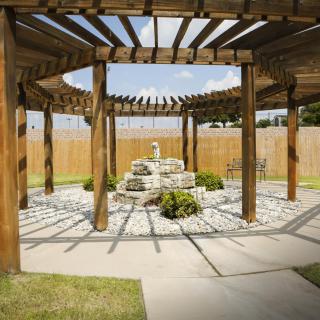 A wooden pergola with a stone fountain at its center, surrounded by a landscaped garden.