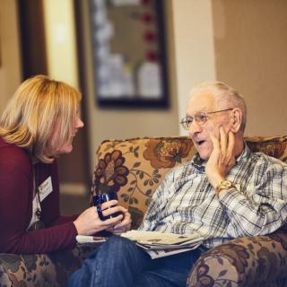A caregiver and an elderly man share a joyful conversation indoors.