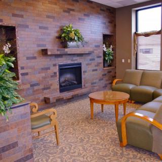 A cozy living room with a modern design featuring a fireplace, green sofas, wooden tables, and decorative plants.