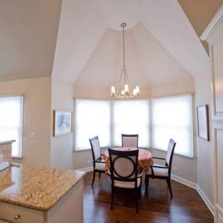 A cozy dining area with a round table, four chairs, and a chandelier, framed by large windows letting in natural light.