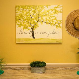 A cozy room with a yellow wall features a tree painting with an inspirational quote, complemented by a straw hat and small plants as decor.