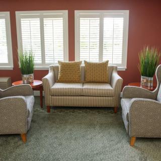 A cozy living room setup featuring a central sofa, two armchairs, side tables, and potted plants against a red wall with large windows.