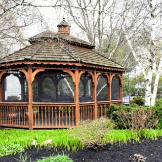 A wooden gazebo in a garden surrounded by trees and greenery.