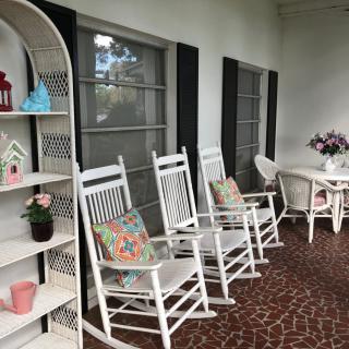A charming porch featuring white rocking chairs with colorful pillows, decorative shelves, and a table adorned with flowers.