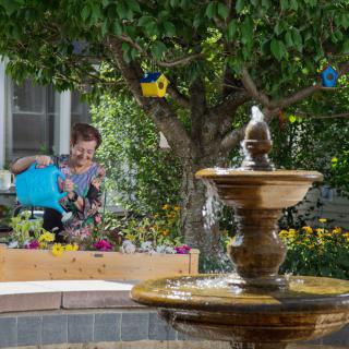 A person waters colorful flowers in a garden near a fountain, with birdhouses hanging from a tree.