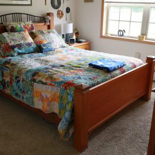 A cozy bedroom with a wooden bed frame and colorful quilt, featuring a window and nightstand.