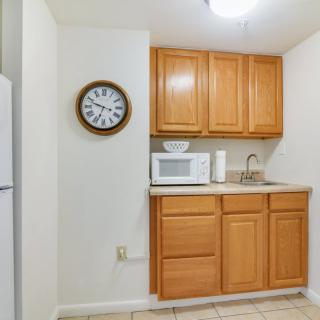 A small kitchen area with wooden cabinets, a microwave, sink, and a clock on the wall.
