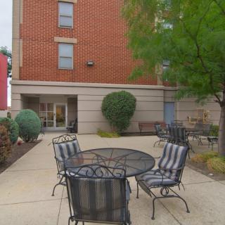 A cozy outdoor patio with metal furniture situated near a brick building, surrounded by trees and landscaping.