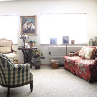 A cozy living room with a mix of floral and plaid patterned furniture, illuminated by natural light from two large windows.