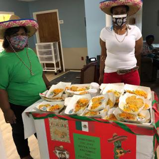 Two women wearing festive hats stand behind a table filled with delicious Mexican food at a community gathering.
