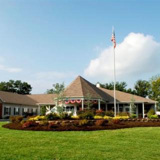 A large building with a sloped roof, surrounded by a well-maintained lawn and gardens, featuring an American flag in the foreground under a clear sky.