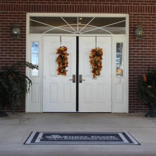 Front entrance of a building with white double doors adorned with autumn decorations and a welcome mat.