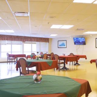A spacious dining area set with green and red tablecloths, featuring multiple tables and chairs, illuminated by natural light.
